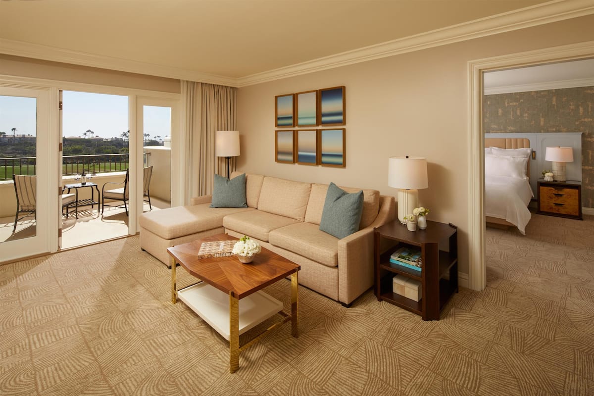 Habitación de hotel con un sofá color beige, una mesa de centro de madera, una mesita auxiliar con una lámpara y vistas al exterior a través de una puerta corrediza.