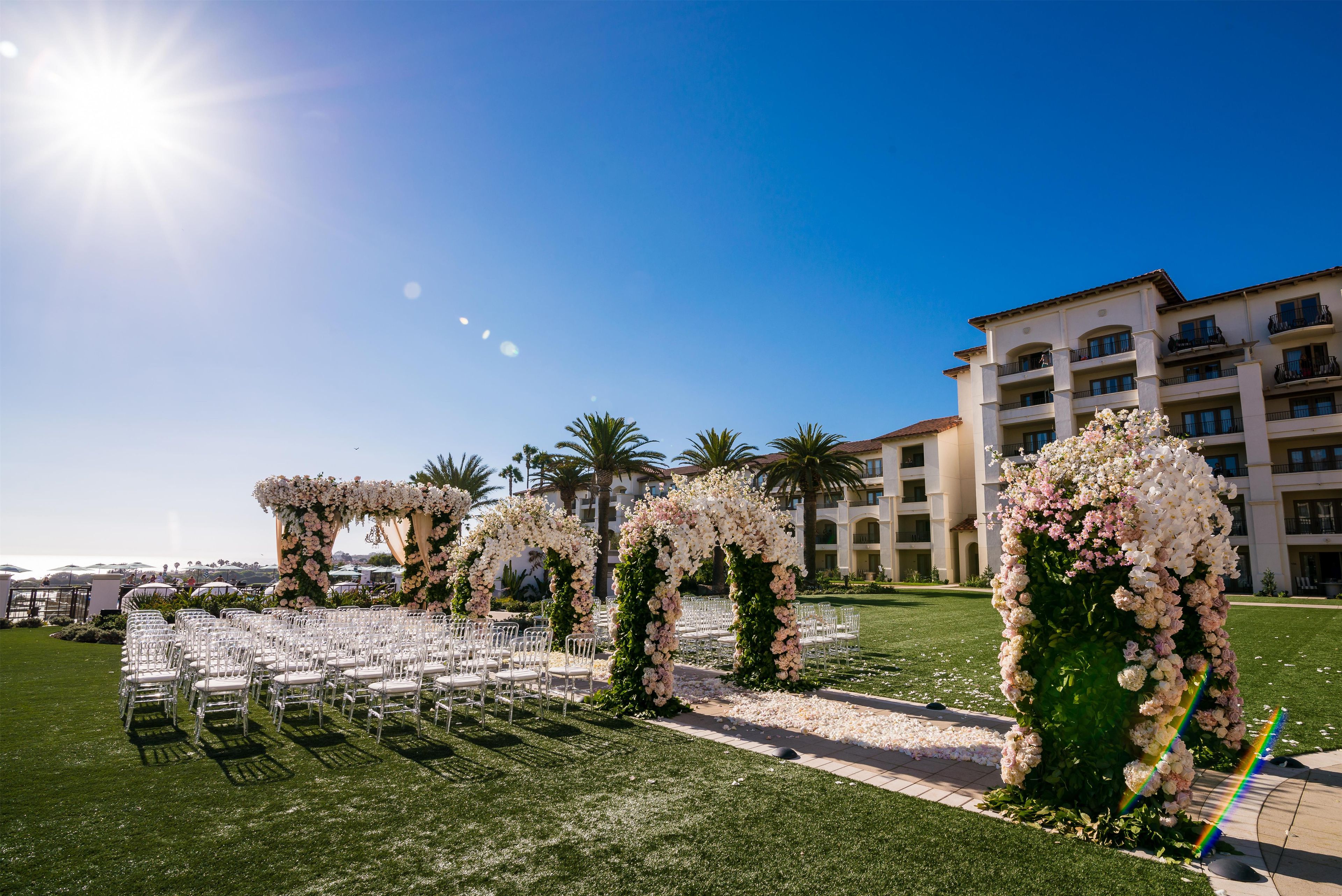 Un espacio para bodas al aire libre con sillas blancas y arcos florales, un edificio al fondo y un cielo despejado.