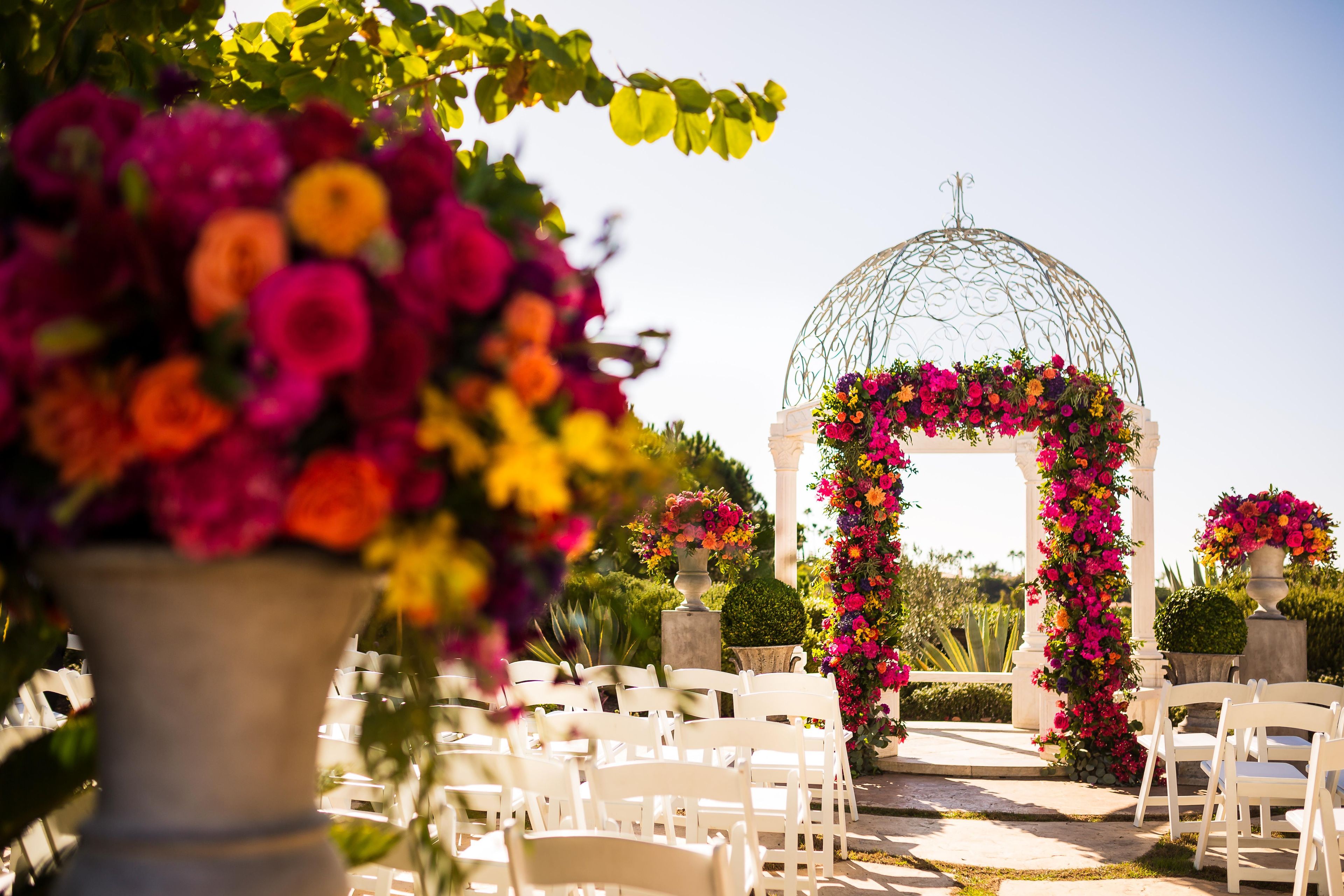 An outdoor wedding venue with a floral archway, white chairs, and a clear sky.
