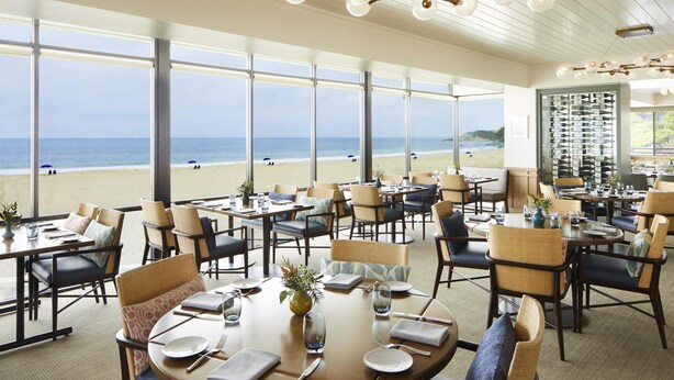 Un restaurant avec des tables et des chaises disposées pour accueillir les clients, de grandes fenêtres donnant sur la plage et un plafond agrémenté d’un luminaire original.