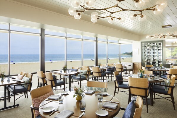 Ein Restaurant mit Tischen und Stühlen, das zum Essen einlädt, große Fenster mit Blick auf den Strand und eine Decke mit einer besonderen Leuchte.