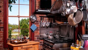 Une cuisine avec un mur en briques, une fenêtre donnant sur un espace verdoyant, une cuisinière et des casseroles suspendues.