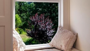 Un siège de fenêtre agrémenté d’un coussin et d’un oreiller, donnant sur un jardin planté d’arbres et d’arbustes.
