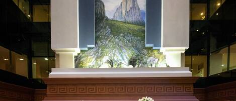 Un vestíbulo con un gran mural de paisaje montañoso, una zona de estar circular con mesas y un pilar decorativo con flores.