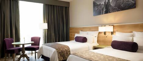 A hotel room with two beds, a table, chairs, a lamp, and a mountain landscape painting on the wall.