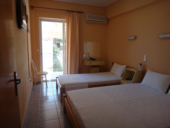 A hotel room with two single beds, a small desk with a chair, a window with curtains, and an air conditioning unit on the wall.
