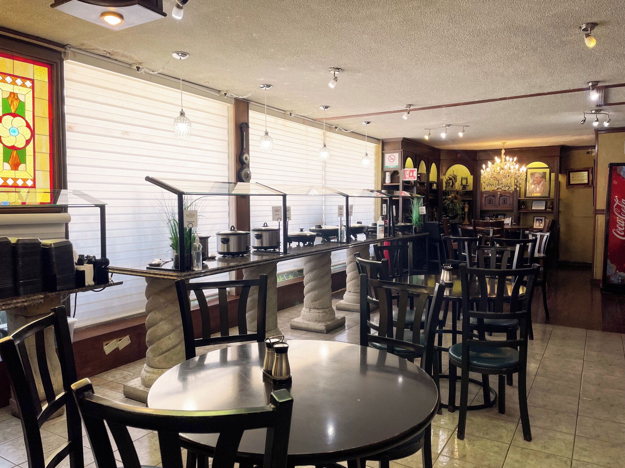 Interior de restaurante con mesas y sillas redondas, una partición de vidrio y un refrigerador Coca-Cola.