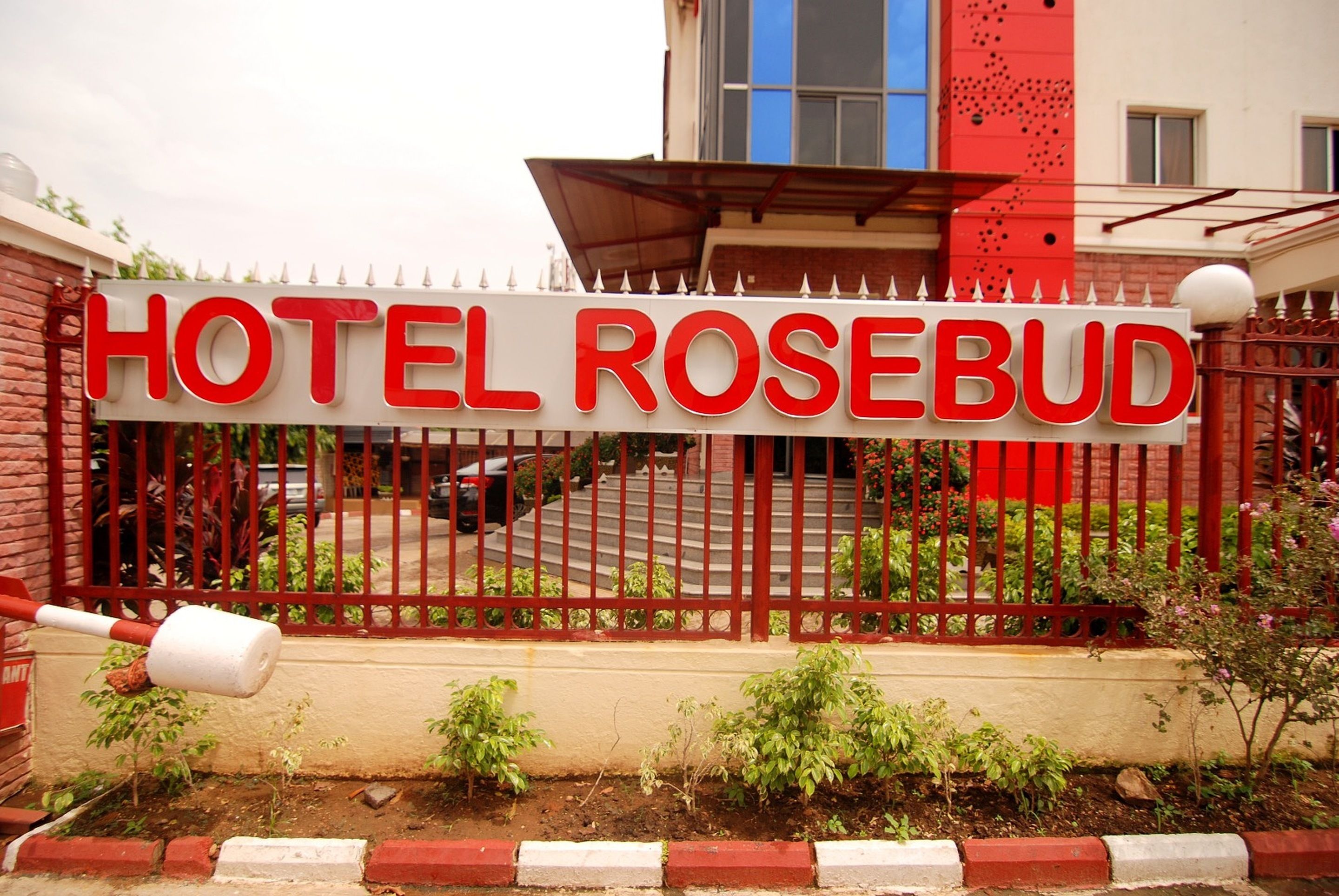 A hotel sign with the name "HOTEL ROSEBUD" in bold red letters on a white background, surrounded by a red metal fence with decorative elements.