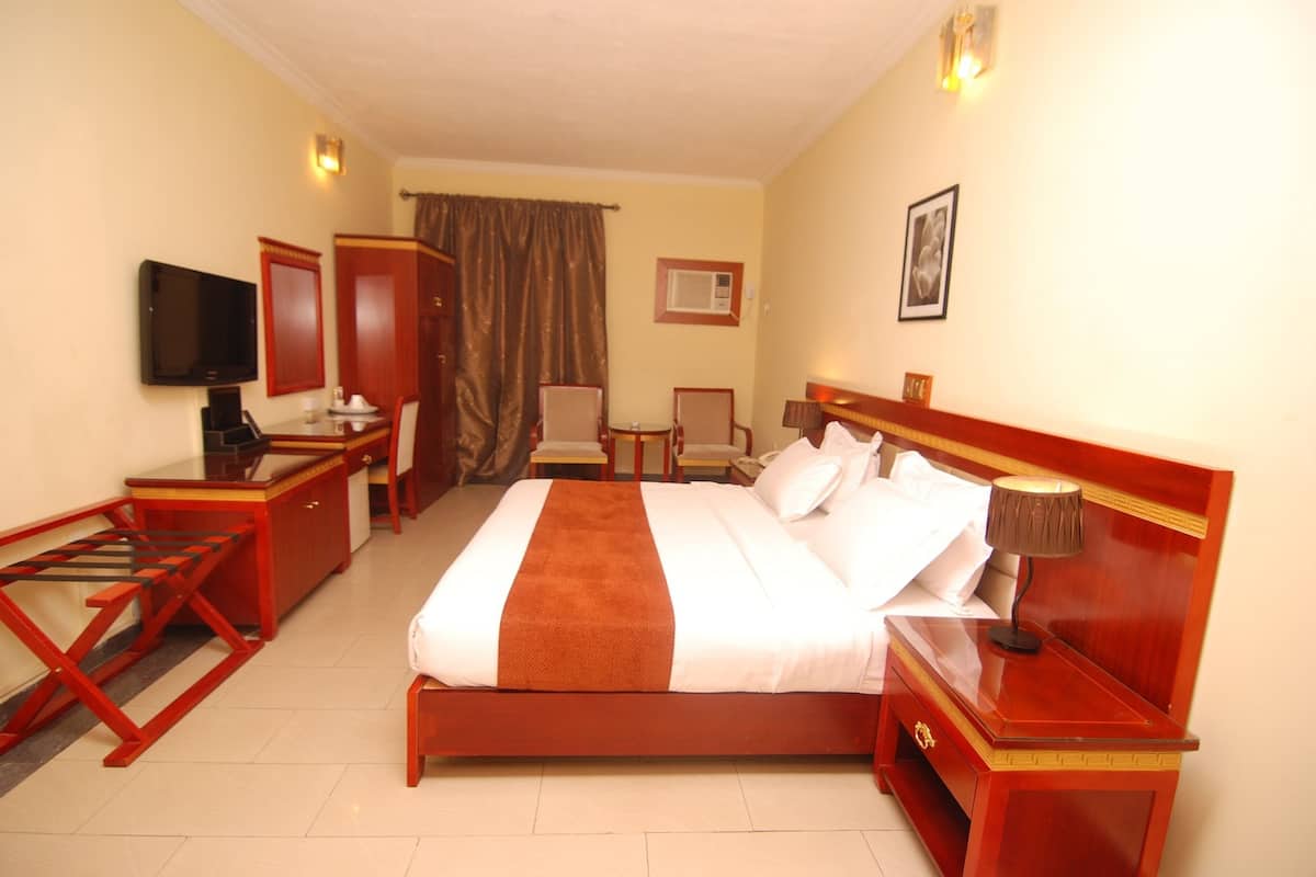 A hotel room with a bed, bedside table, lamp, TV, wardrobe, and a desk with chairs.