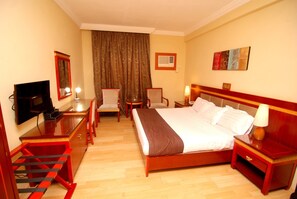 A hotel room with a bed, a desk with a TV, a chair, a lamp, and a painting on the wall. - Hotel Rosebud (Abuja)