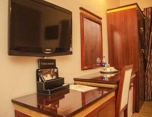 A hotel room with a flat-screen TV mounted on the wall, a wooden cabinet, a desk with a chair, and a small table with a bottle of water and a cup. - Hotel Rosebud (Abuja)