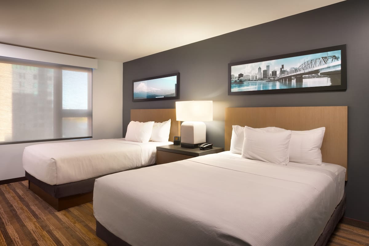 A hotel room with two beds, a large window, a cityscape painting, and a television on the wall.