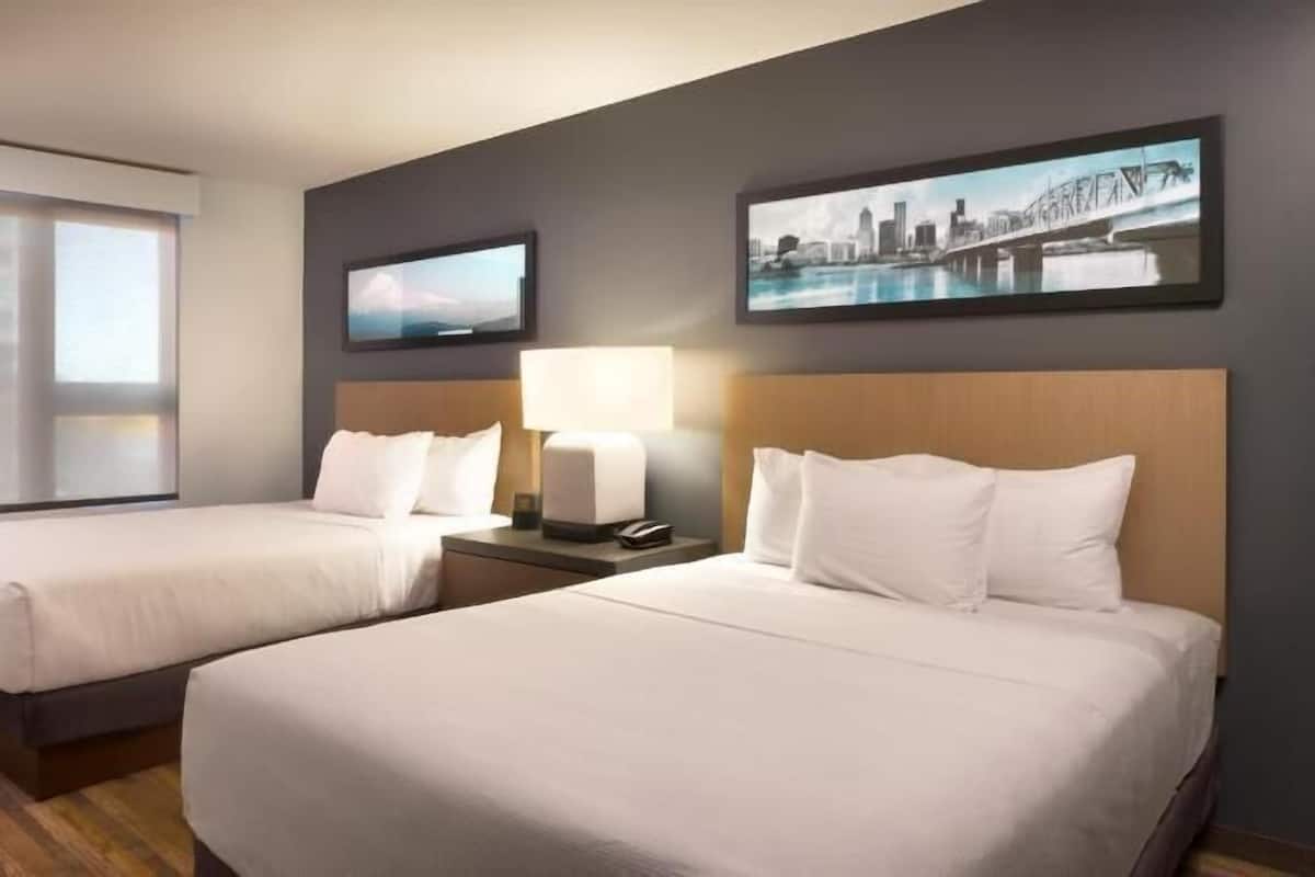 A hotel room with two beds, a window, a nightstand with a lamp, and two wall art pieces.