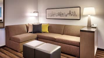 A modern hotel room with a sectional sofa, a small ottoman, a bedside table with a lamp, and a framed cityscape artwork on the wall.