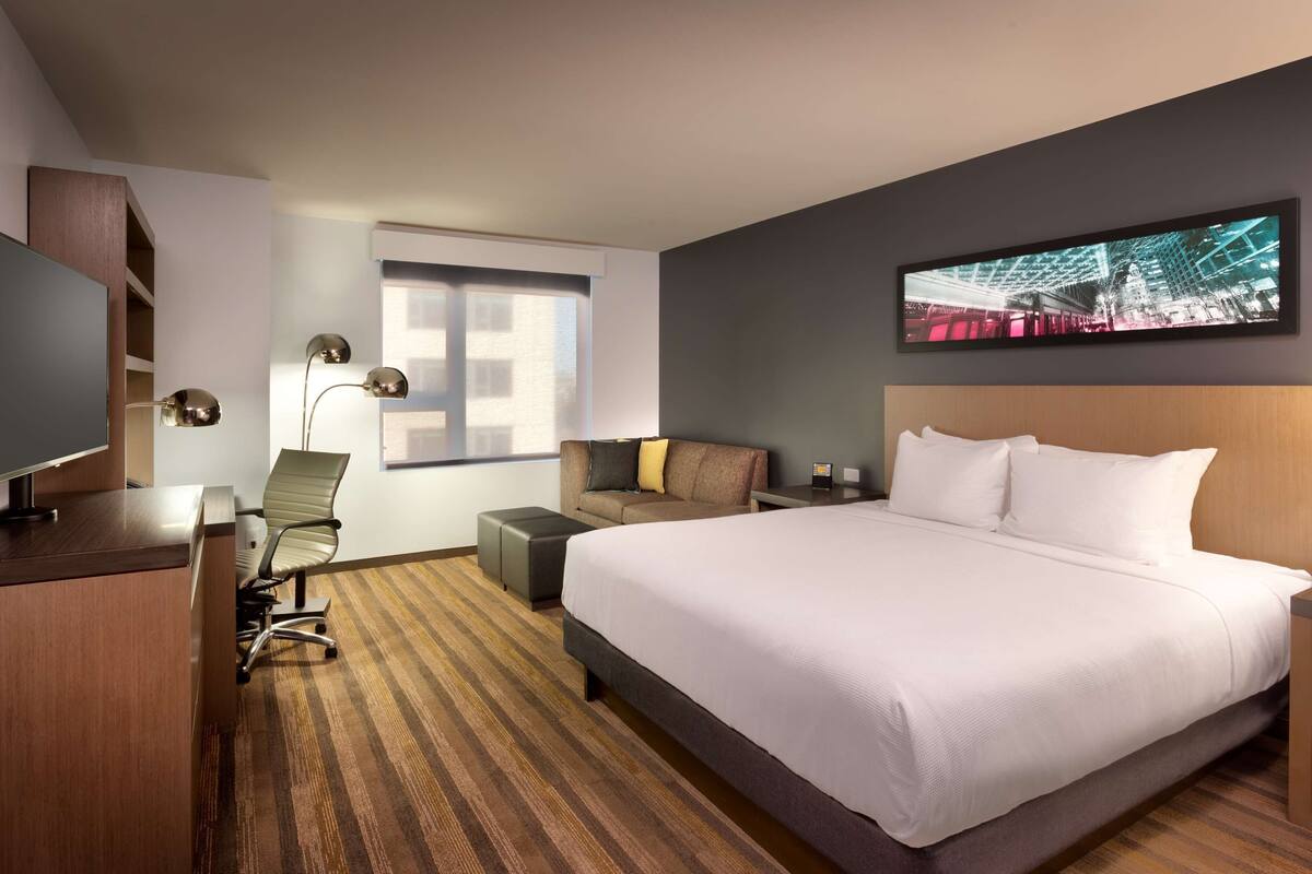 A hotel room with a large bed, a desk with a computer, a chair, a sofa, and a TV.