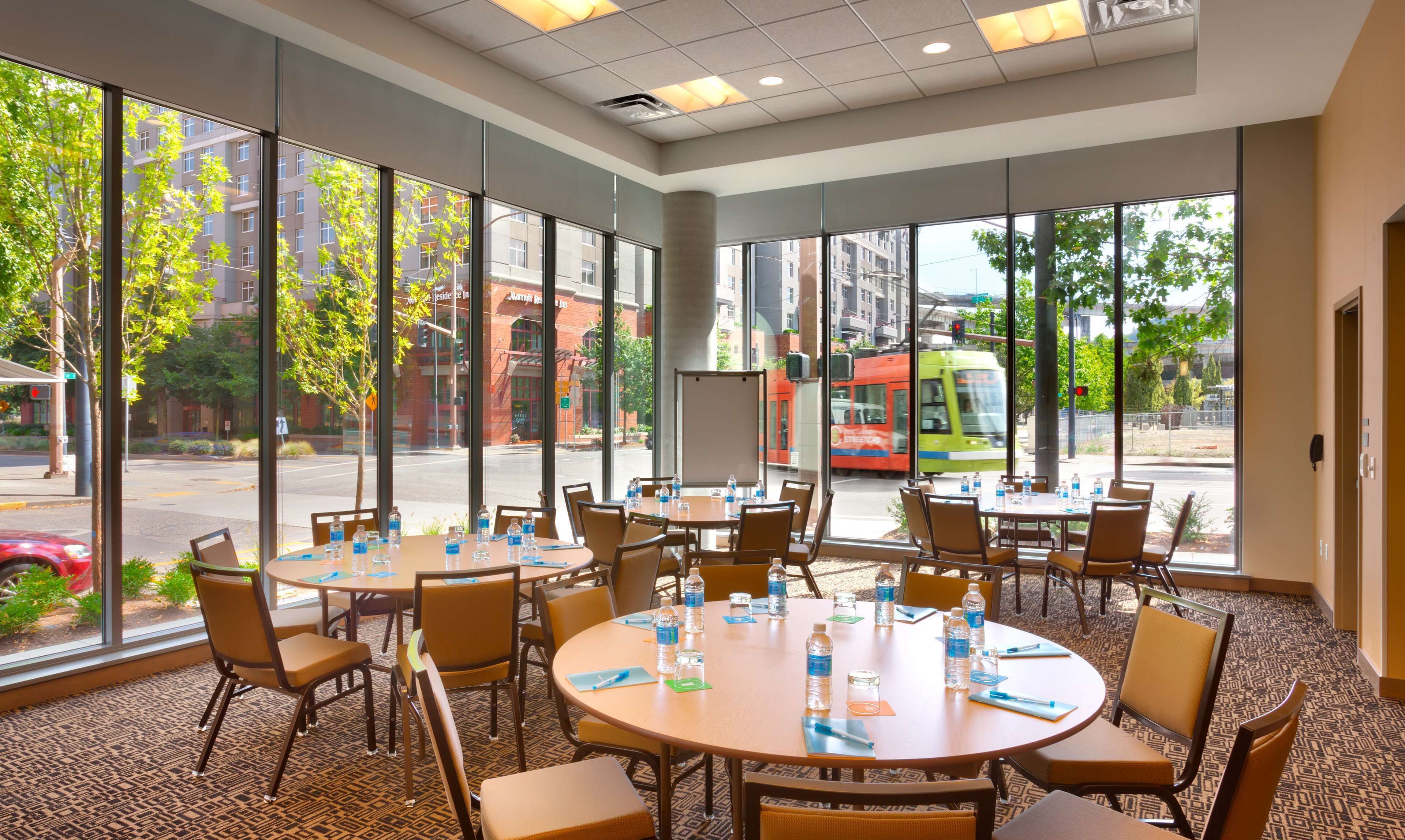 A conference room with large windows, round tables, and chairs, equipped with water bottles and notepads.