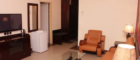 A hotel room with a flat-screen TV, a sofa, a coffee table, a small white refrigerator, a mirror, and an air conditioning unit.