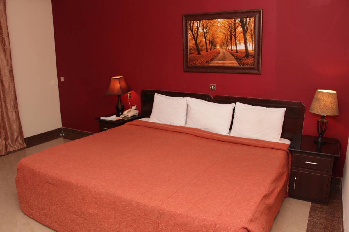 Une chambre d’hôtel avec un grand lit, des murs rouges, un tableau encadré au mur et deux tables de chevet avec des lampes.