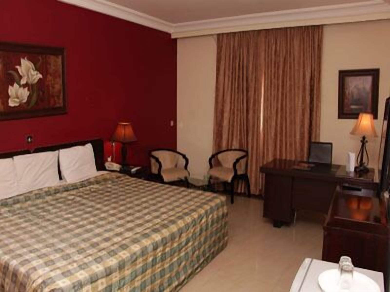 A hotel room with a checkered bedspread, a desk, two chairs, a lamp, and a painting on the wall.