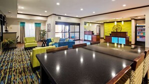 Un vestíbulo de hotel con una amplia mesa de conferencias, zona de estar con sillas, mostrador de recepción y un refrigerador.