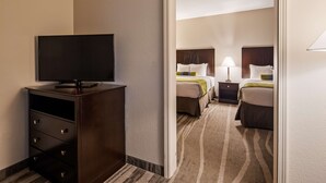 Una habitación de hotel con una televisión sobre un mueble de madera, dos camas con cabeceras, una mesita de noche con una lámpara y una alfombra a rayas.