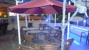 Un patio al aire libre con mesas redondas y sillas de mimbre, un paraguas grande y una piscina con iluminación azul.