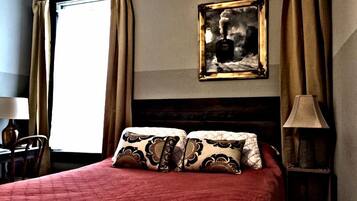 Habitación de hotel con una cama grande, cabecera, mesita de noche con lámpara, ventana con cortinas y un cuadro en la pared.
