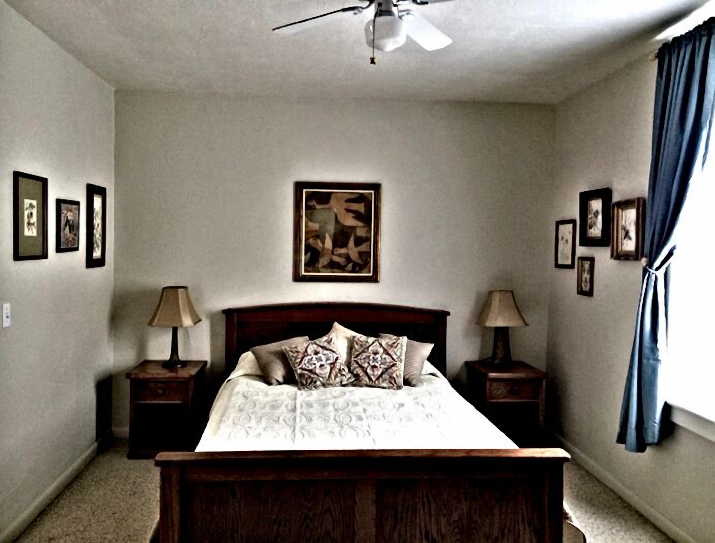 A bedroom with a bed, two nightstands, a ceiling fan, and framed pictures on the walls.