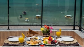 A breakfast table with plates of food, a fruit bowl, and a vase of flowers, overlooking a beach with boats. - Pousada dos Jangadeiros (Maragogi)
