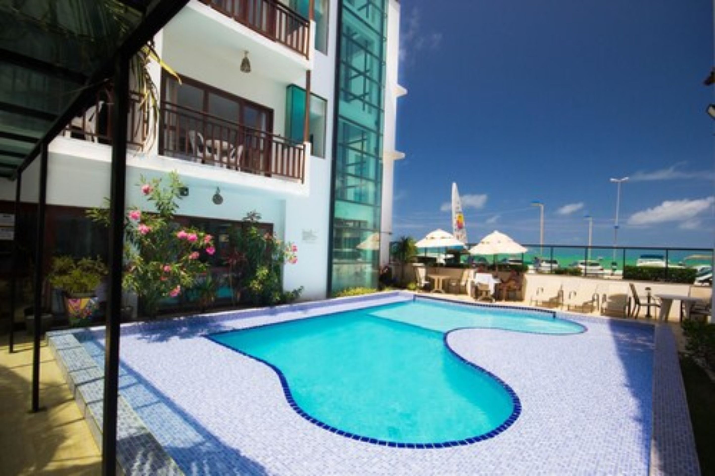 A swimming pool with a tiled deck, surrounded by a building with balconies and a view of the beach.