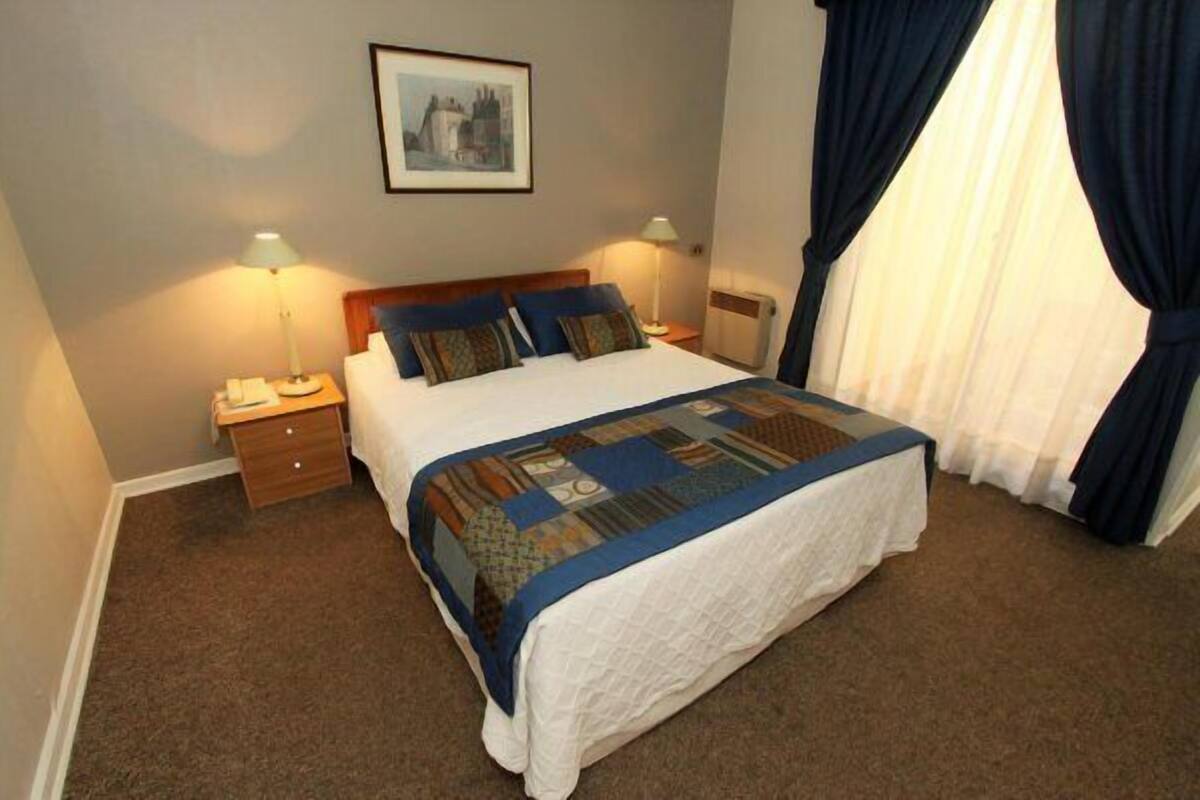 A neatly made bed with a quilt, two bedside lamps, a nightstand, and a framed picture on the wall.