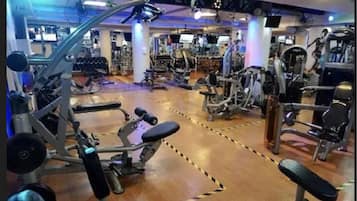 Academia com diversos equipamentos de exercício, piso de madeira e várias telas de TV nas paredes.