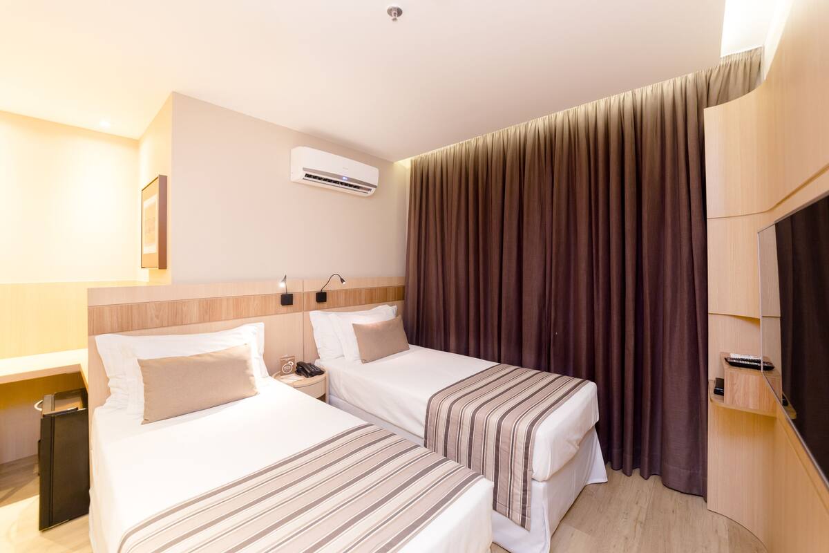 A hotel room with two beds, a bedside table with a lamp, an air conditioning unit, and a large window with curtains.