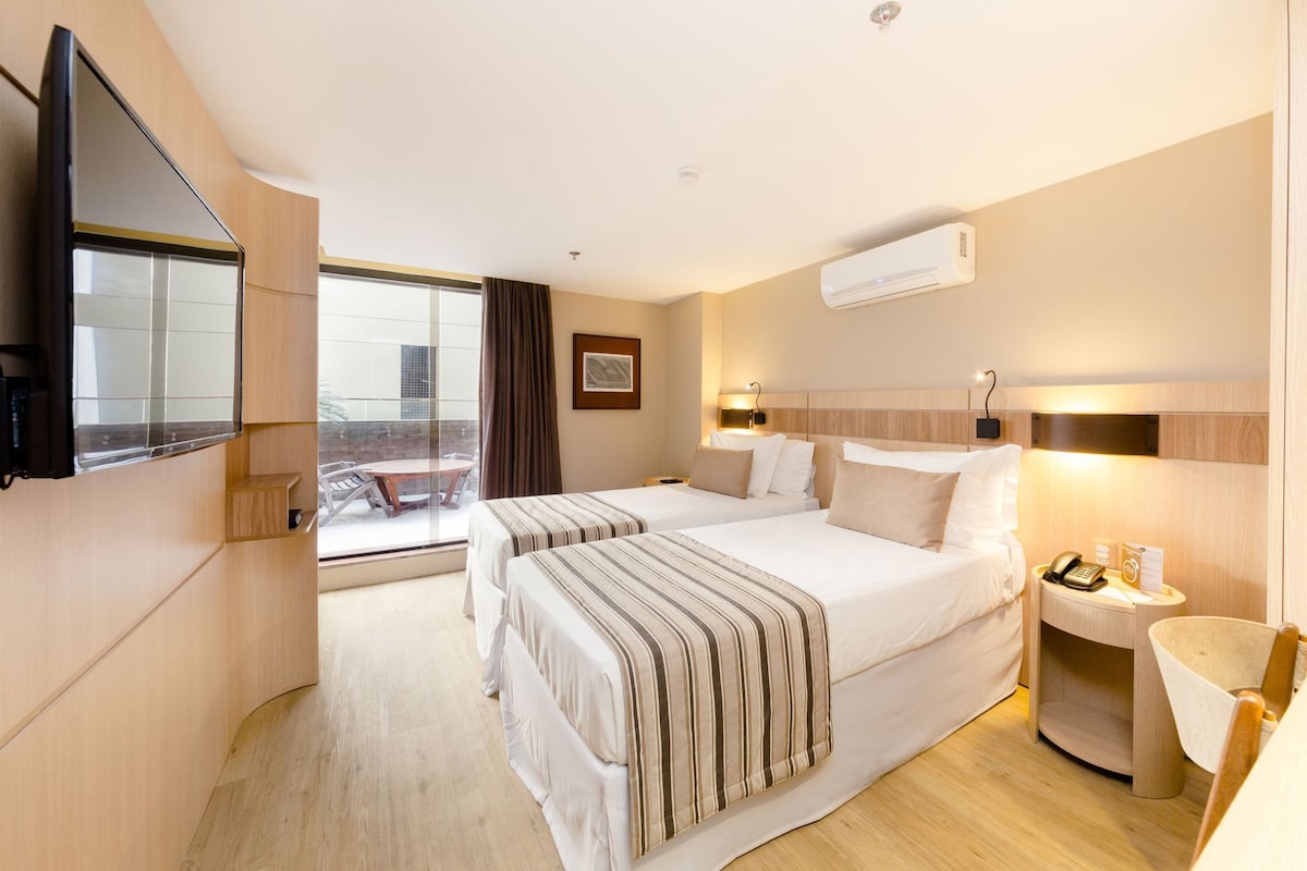 A hotel room with a large bed, a TV mounted on the wall, a bedside table with a lamp, and a window with curtains.