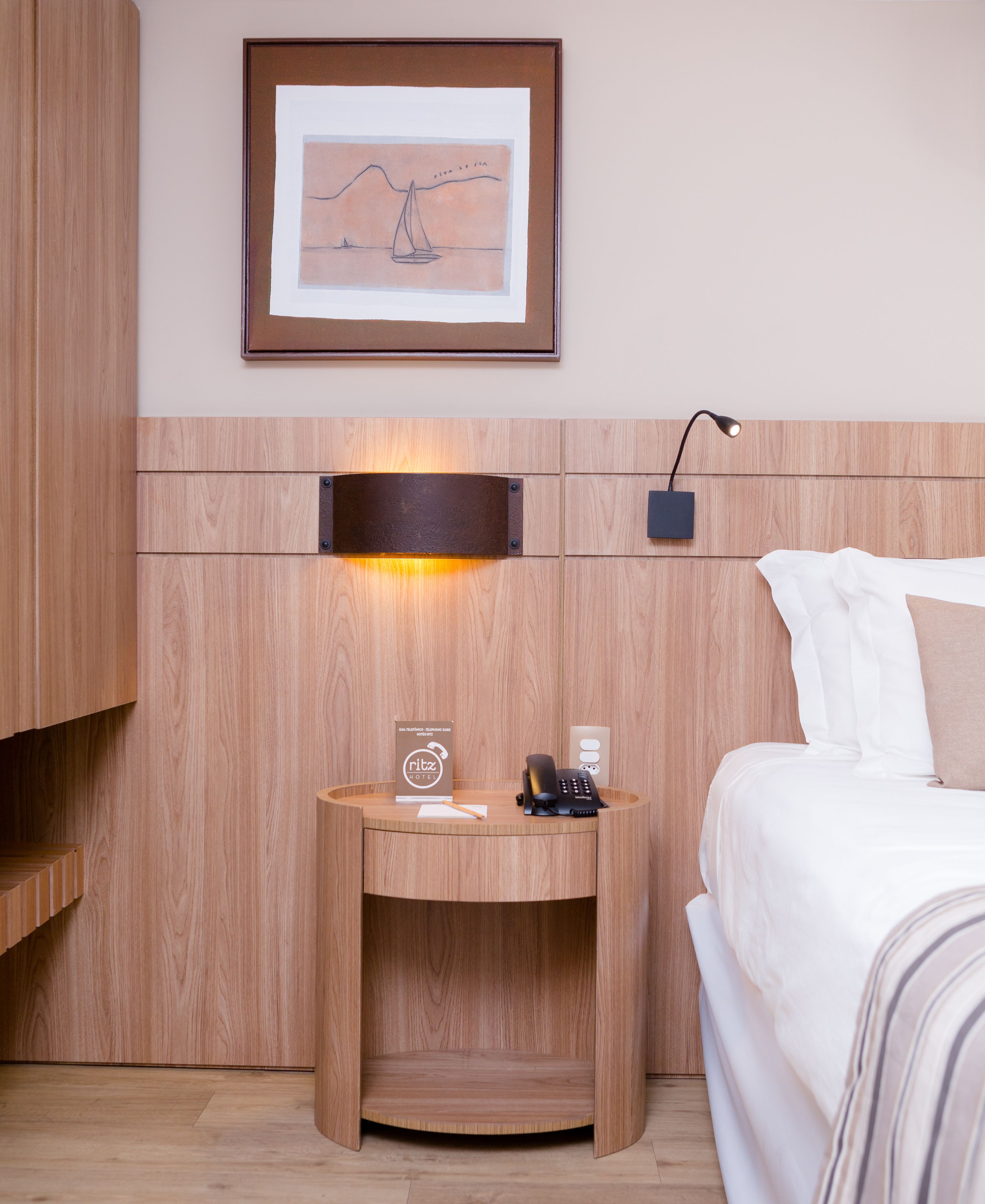 A hotel room with a wooden headboard, a framed picture above the bed, a bedside table with a phone and a sign, and a lamp on the wall.