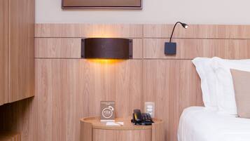 A hotel room with a wooden headboard, a framed picture above the bed, a bedside table with a phone and a sign, and a lamp on the wall.
