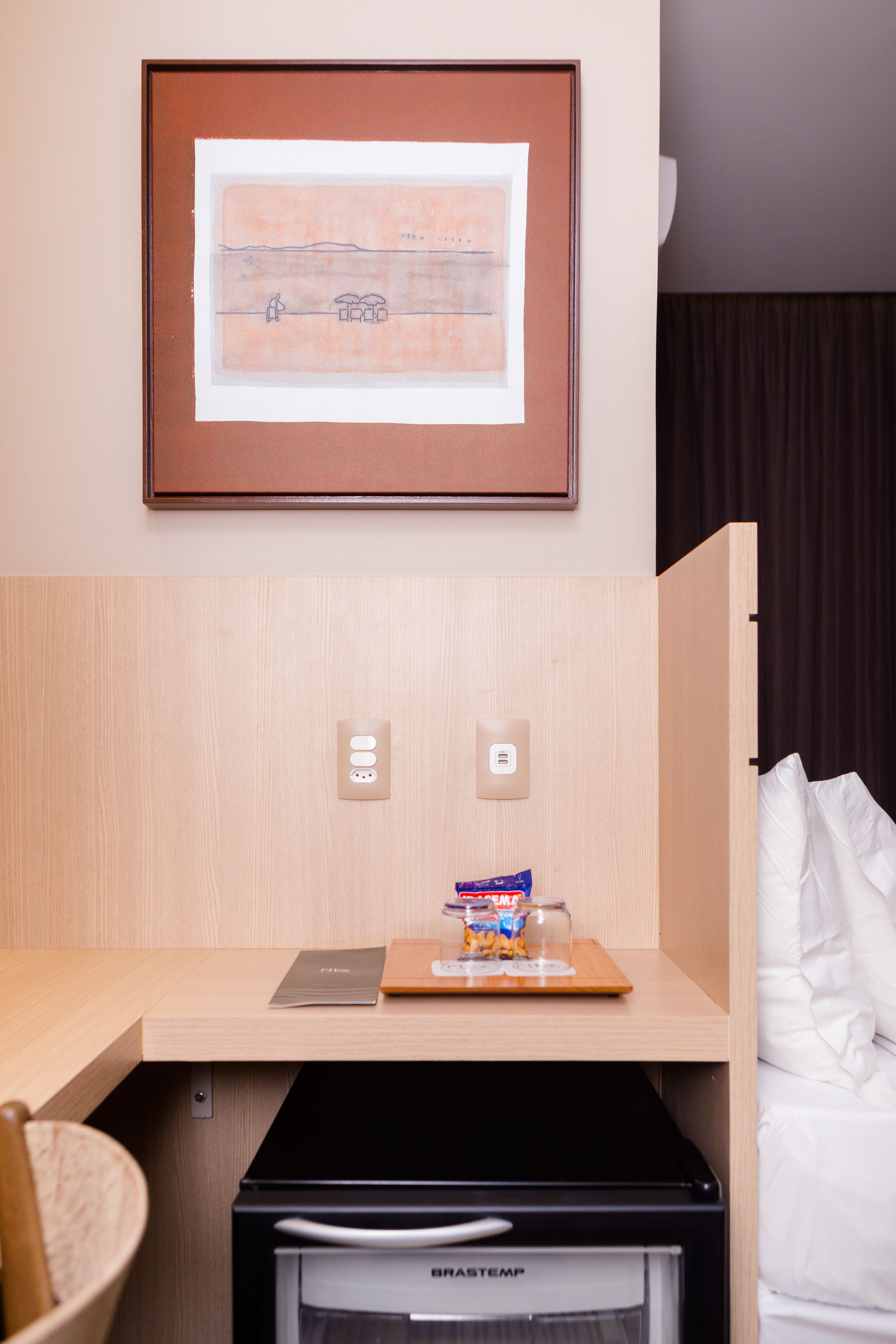 A compact hotel room with a wooden desk, a small refrigerator, a bed with white linens, a framed picture on the wall, and a power outlet.