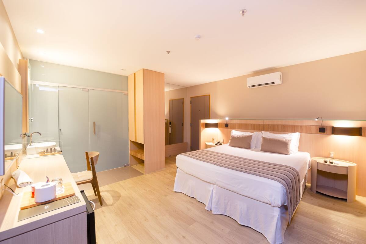 A modern hotel room with a large bed, bedside tables, a mirror, a wardrobe, and a bathroom with a sink and toilet.