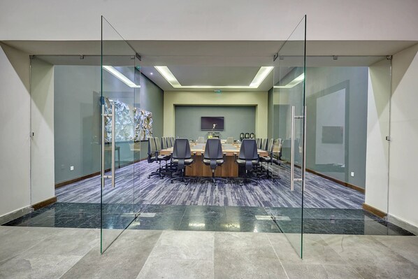 Un salón de conferencias con un panel de vidrio, una mesa larga con sillas y un televisor montado.