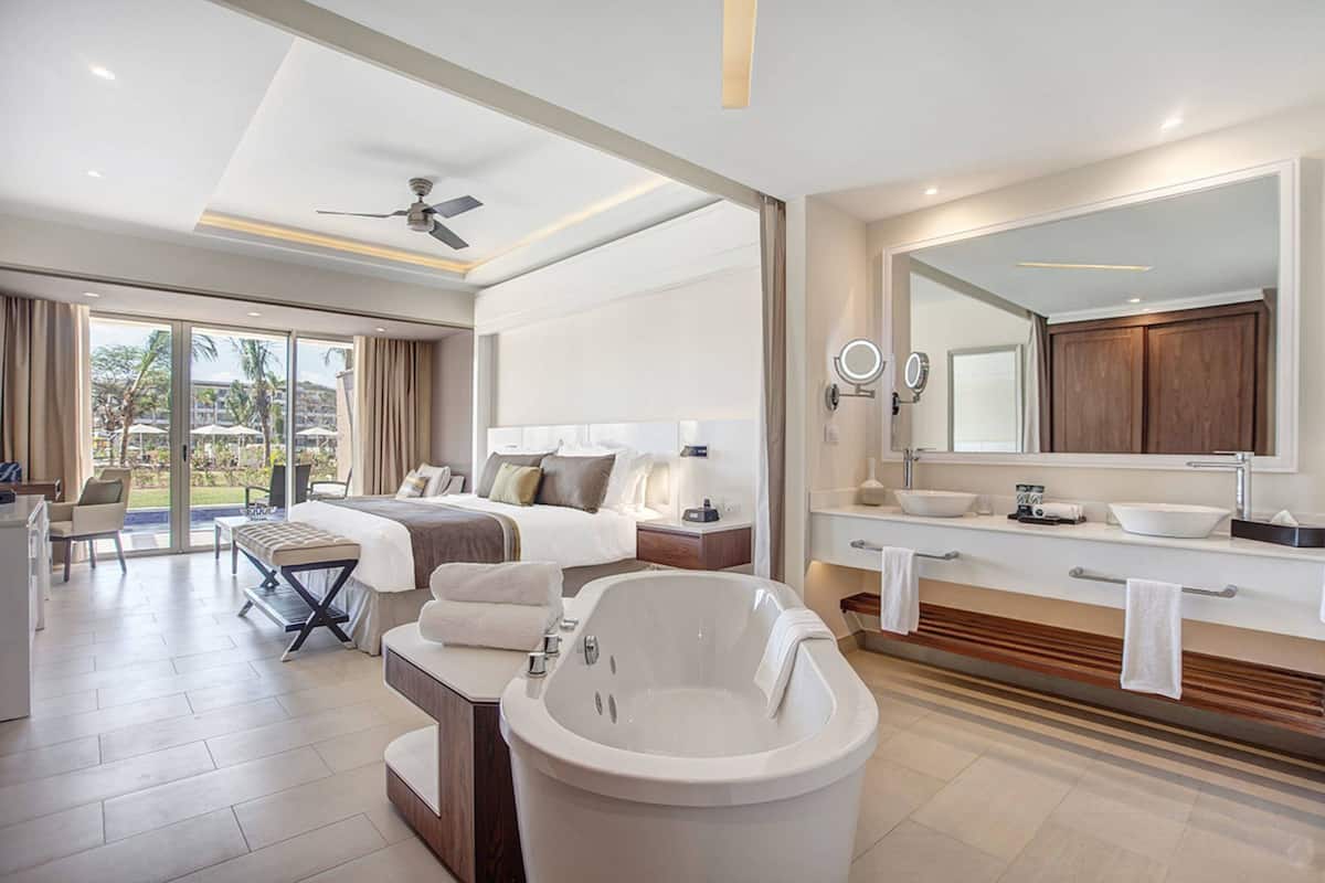 Ein modernes Hotelzimmer mit einem großen Bett, einer Badewanne, einer Sitzecke mit Tisch und einem Balkon mit Blick auf Palmen.