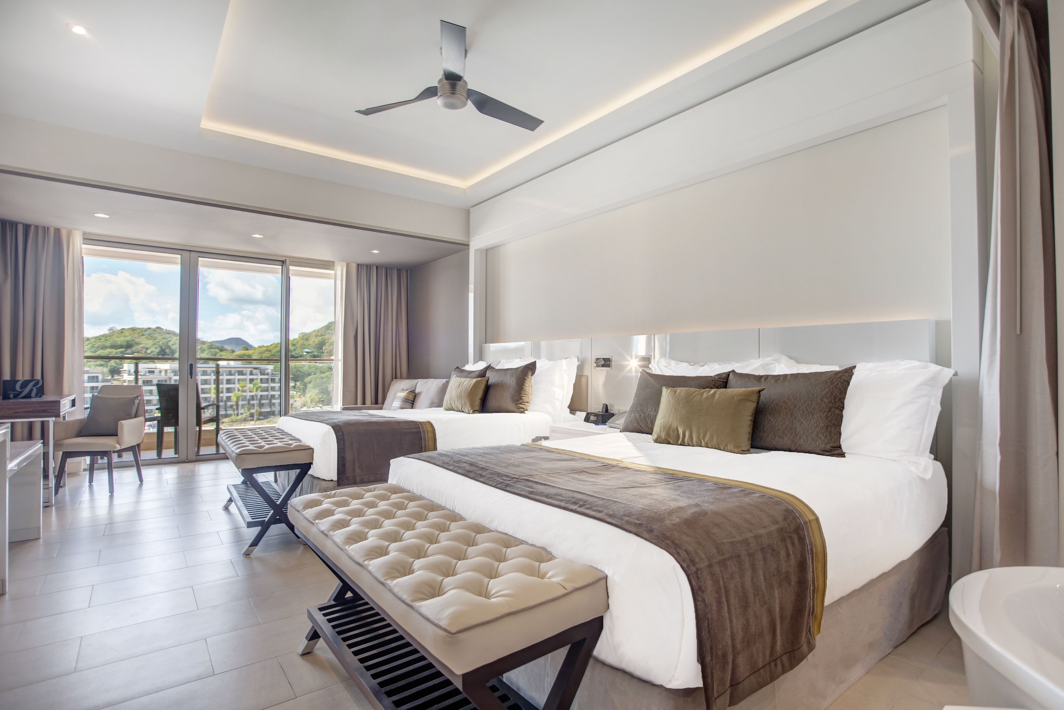 A modern hotel room with a large bed, a bench, a desk, and a view of the outdoors.