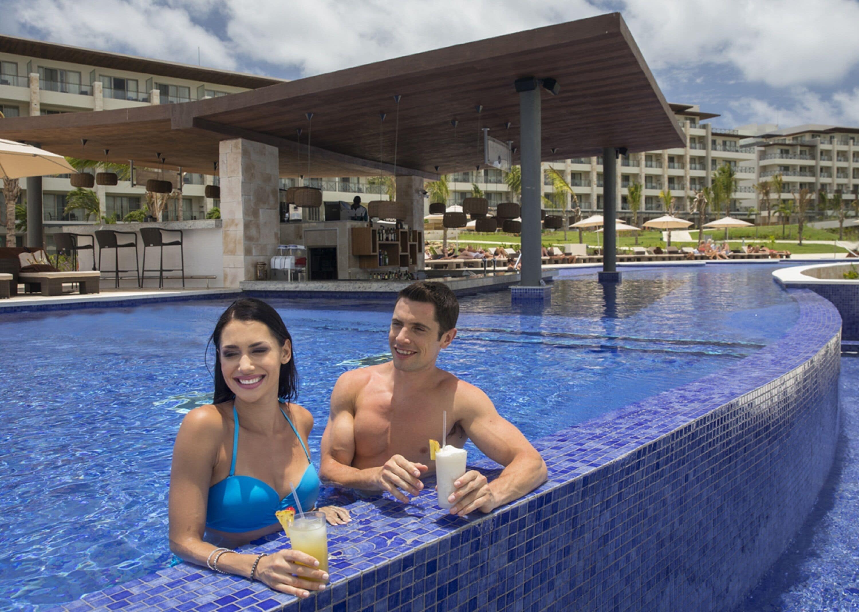 Una pareja en una piscina con borde de azulejos, sillas de descanso y una zona cubierta al fondo.