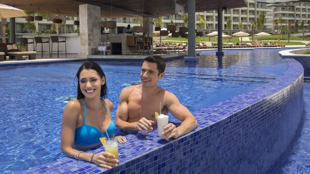 A couple in a pool with a tiled edge, lounge chairs, and a covered area in the background.
