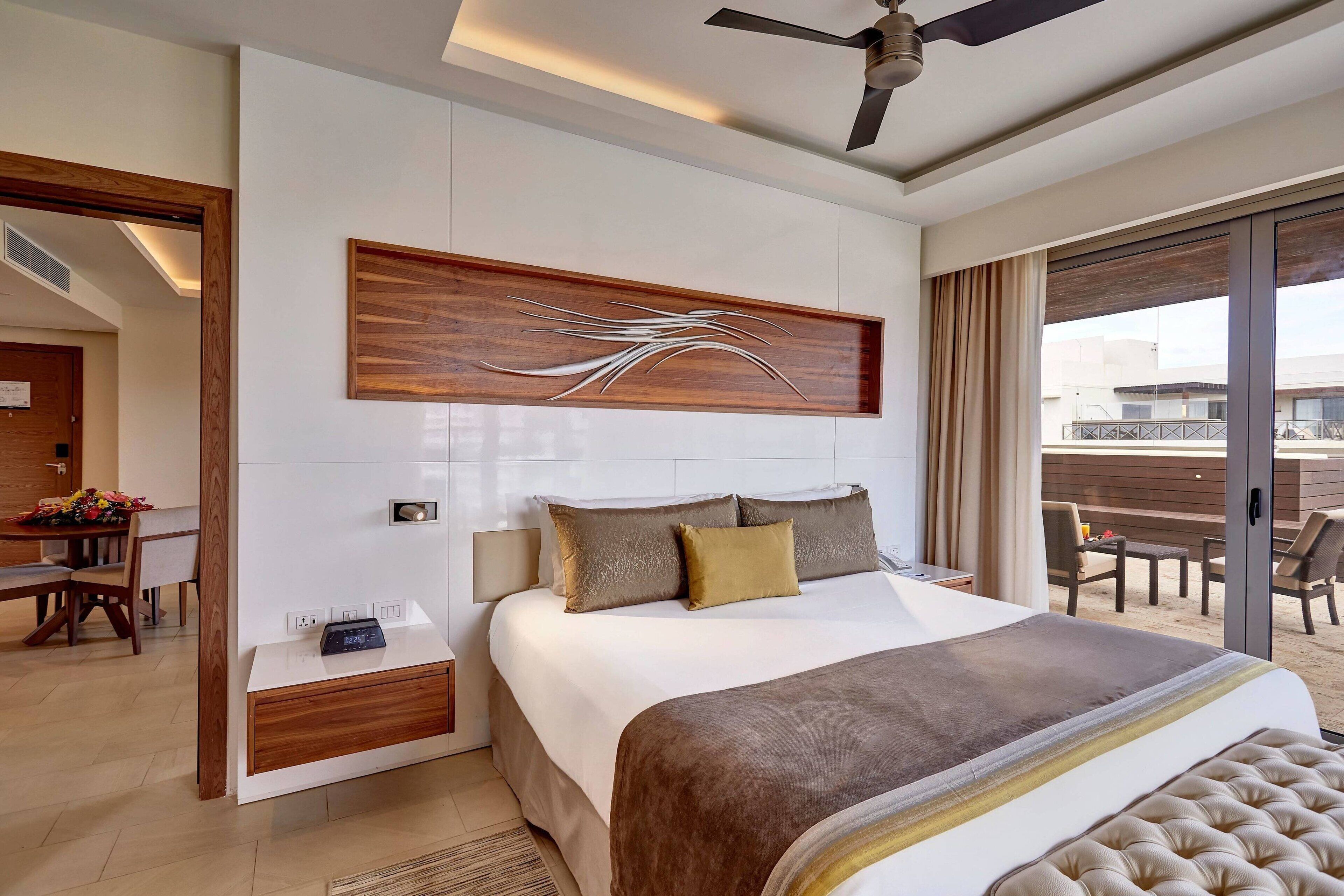 A modern hotel room with a large bed, a ceiling fan, a wooden accent wall, and a view of the outdoors.