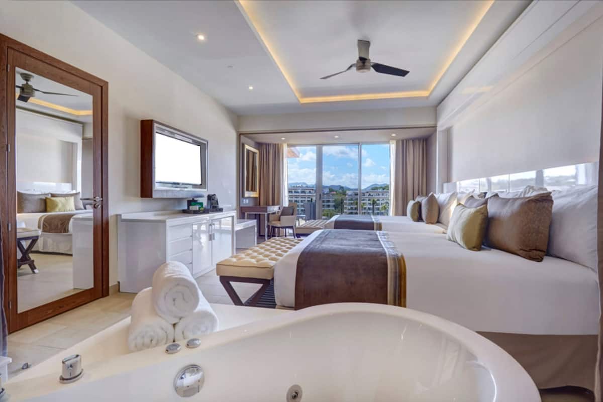 Ein Hotelzimmer mit einem großen Bett, einer Schlafcouch, einem Schreibtisch mit Stuhl, einem großen Fenster mit Blick auf die Stadt, einem Deckenventilator und einer Badewanne.