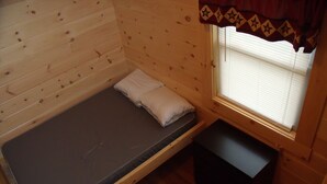 A small, single-bed room with a wooden floor and walls, a window with blinds, and a small black storage bench. - Shenandoah Valley Campground (Quicksburg)