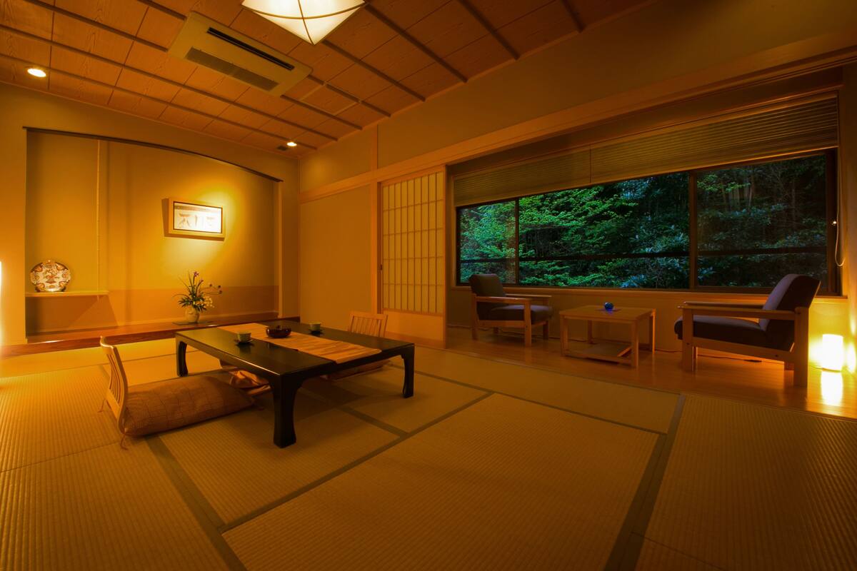 A traditional Japanese room with tatami flooring, a low table, chairs, and a large window overlooking a forest.