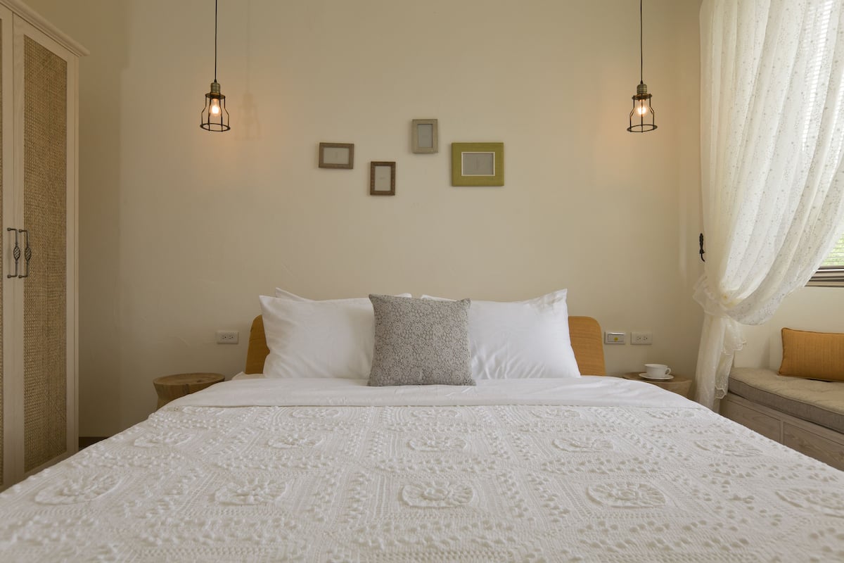 A neatly made bed with a white bedspread, a gray pillow, and a wooden headboard. There are two bedside tables with a cup and saucer on one, and a small wooden stool on the other. The room has a simple, clean design with a plain wall and a window with sheer curtains.
