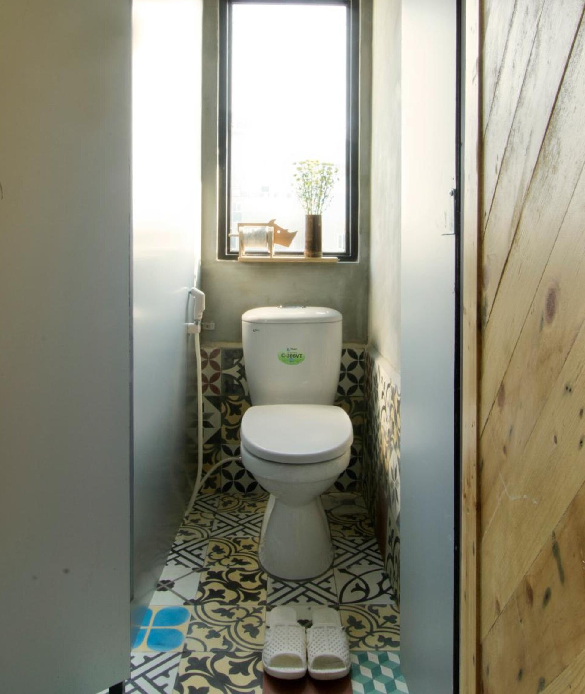 A small bathroom with a toilet, a window with a plant, and a pair of slippers on the floor.