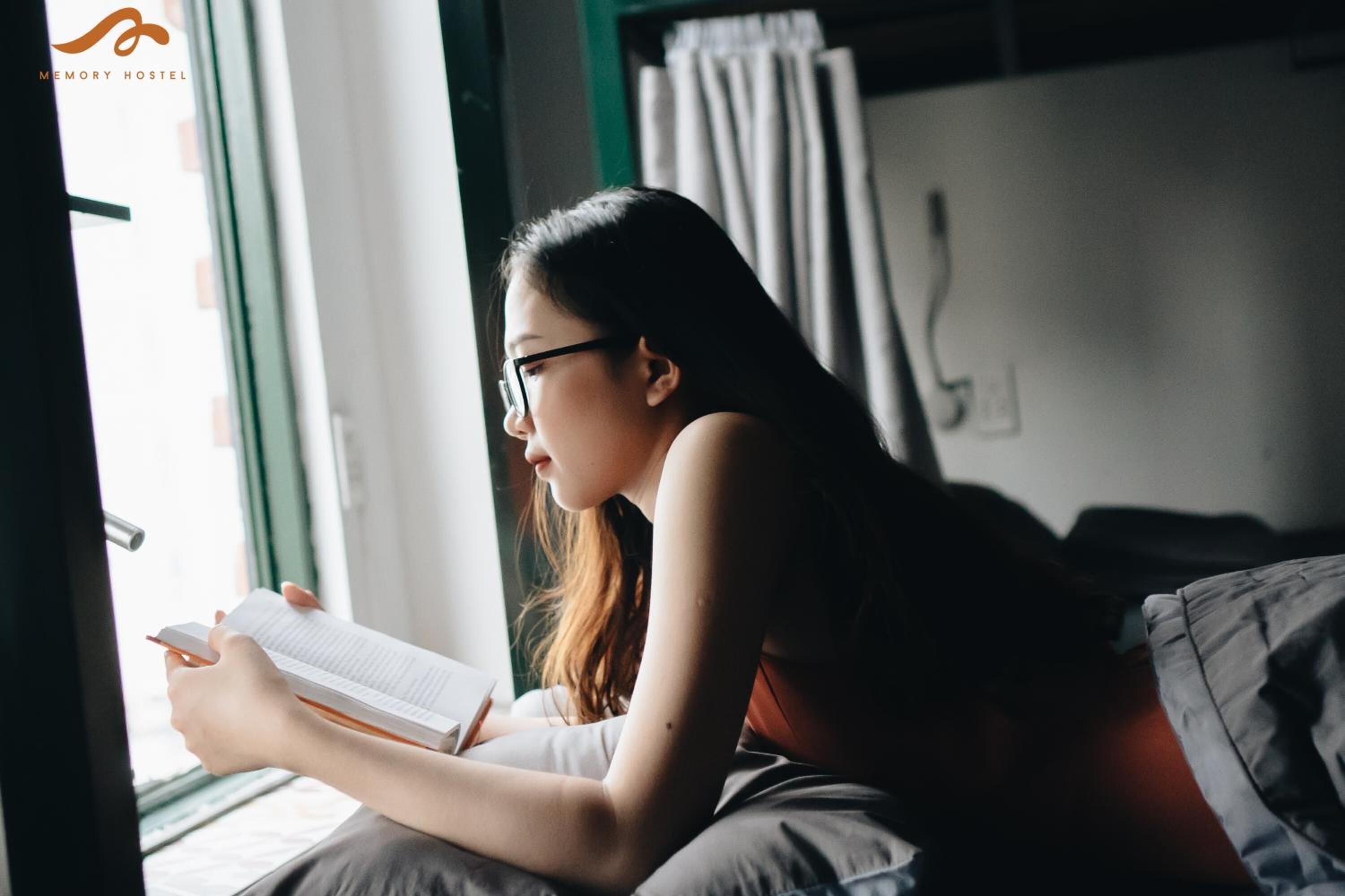 A person reading a book in a room with a window, a bed, and a curtain.